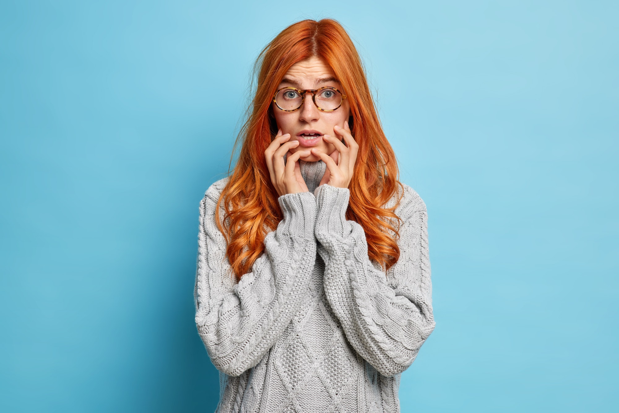 Puzzled nervous woman with long red hair keeps hands on face looks with worried expression at camera