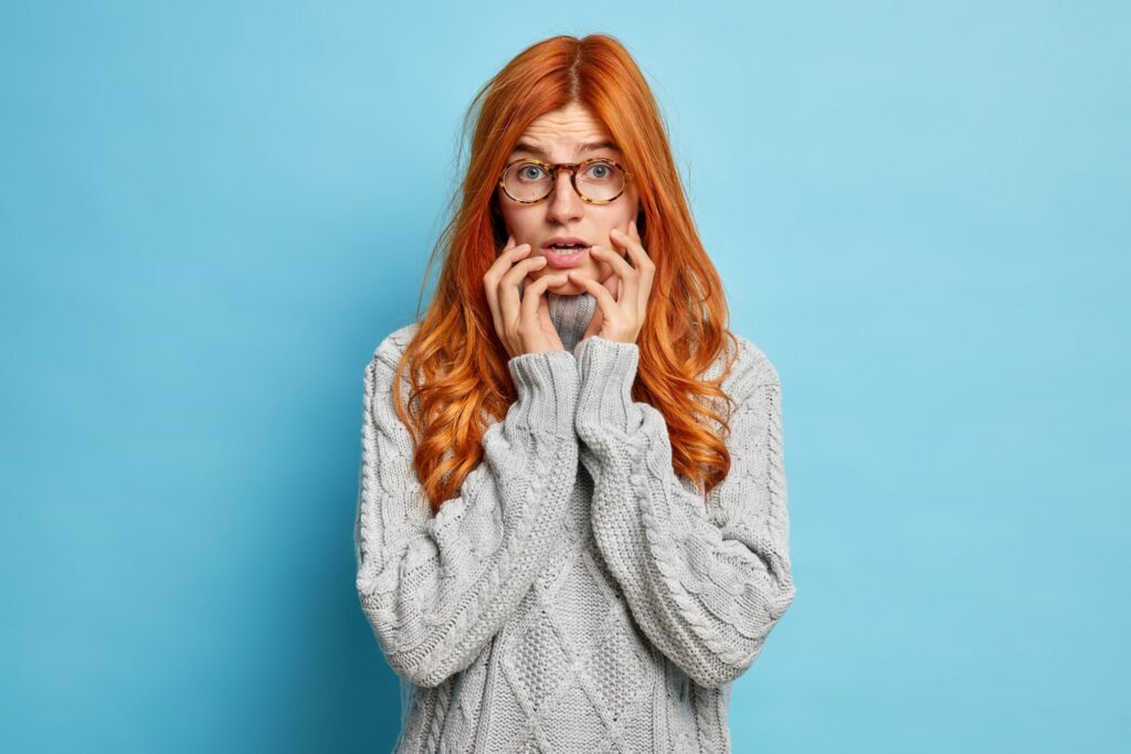 Puzzled nervous woman with long red hair keeps hands on face looks with worried expression at camera